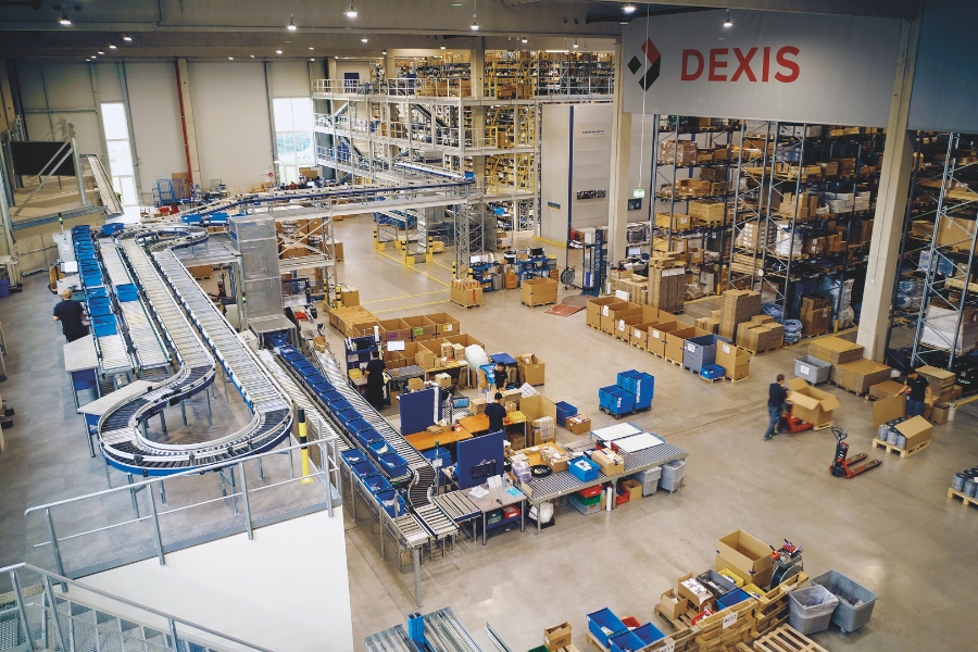 Ein geräumiges Lager mit mehreren Förderbändern, die Pakete transportieren, zeigt das technische Sortiment von DEXIS Austria. Zahlreiche Regale sind mit verschiedenen Artikeln bestückt, und Mitarbeiter verwalten die Abläufe an verschiedenen Stationen.